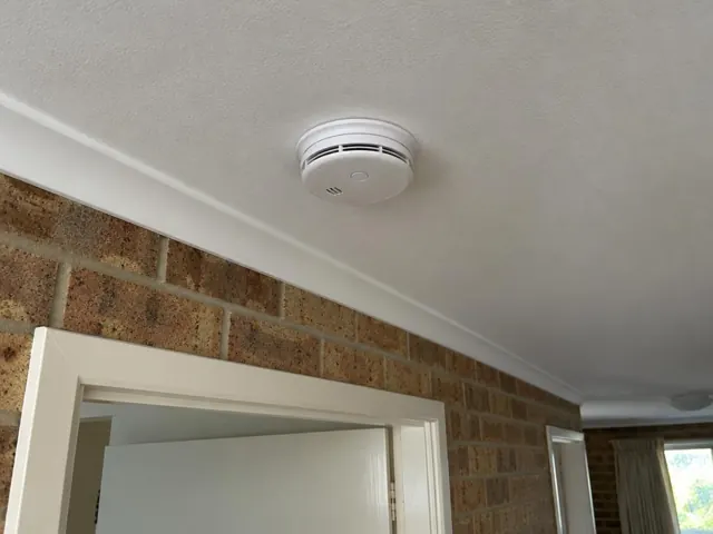 Smoke alarm mounted on a white ceiling above a brick hallway in Noraville